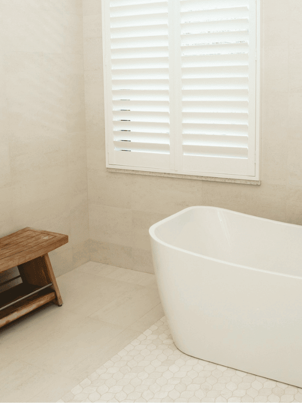 Modern freestanding bathtub in a minimalist bathroom with neutral tones, natural light from plantation shutters, and a sleek wooden stool for a calming, spa-inspired design.