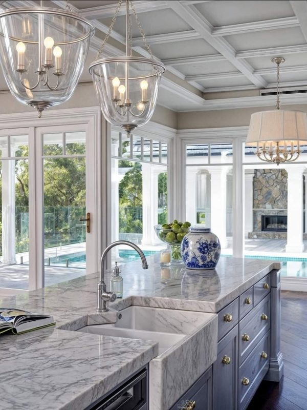 Elegant kitchen featuring a marble island, chandelier lighting, and large windows with a view of the pool and outdoor living space, designed by Aurea Kitchens.