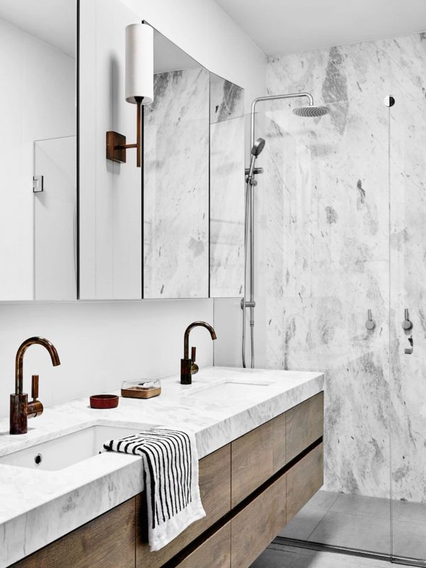 Sleek bathroom vanity with marble countertop and bronze fixtures, featuring a double sink, modern cabinetry, and a walk-in shower with marble walls, showcasing contemporary bathroom design by Aurea Kitchens.
